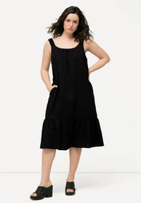 Schwarzes ärmelloses Kleid mit lockerer Passform, gerafft am Hals und mit Rüsche am Saum. Es hat seitliche Taschen und besteht aus leichtem Stoff.