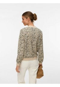 Femme aux cheveux en chignon, portant un pull beige à taches noires et un pantalon blanc, tenant un petit sac marron dans la main droite.