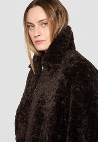 Manteau en peluche marron avec un col haut et une texture douce. Le design est surdimensionné avec une coupe décontractée et des accents en métal.