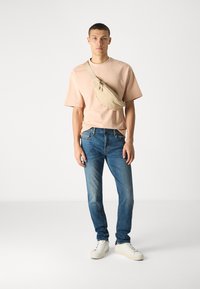 Mlad moški stoji, oblečen v breskevasto majico, modre kavbojke, bele superge in bež torbico čez ramo na belem ozadju.