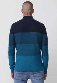 Pletena jopica z visokim ovratnikom, črtastim dizajnom v mornarsko modri, teal in svetlejši modri barvi, z rebrastimi manšetami in robom.
