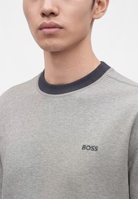 Homme portant un pull gris clair à col rond avec un col gris foncé et un petit logo noir "BOSS" sur la poitrine, sur un fond uni.