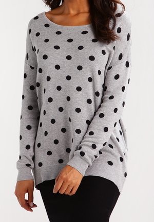 Kvinde iført en grå langærmet trøje med sorte polkaprikker og ribbet kant, kombineret med sorte bukser, stående foran en hvid baggrund.