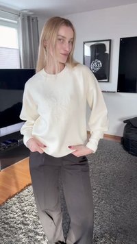 Νέα γυναίκα με μακριά ξανθά μαλλιά ποζάρει σε εσωτερικό χώρο φορώντας ένα εκρού πουλόβερ και σκούρο παντελόνι σε ένα μοντέρνο σαλόνι.
