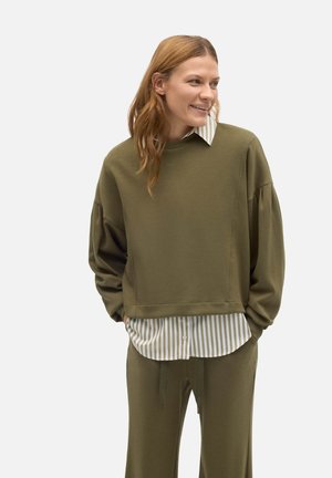 Sudadera verde oliva de corte holgado, fabricada con un tejido texturado y con hombros caídos, llevada sobre una camisa de botones a rayas blancas y grises.