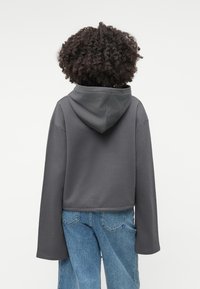 Γκρι κοντό φούτερ με μεγάλη, ρυθμιζόμενη κουκούλα και φαρδιές, μακριές μανίκες. Φοριέται με ανοιχτόχρωμα τζιν, που διαθέτουν κλασικό σχέδιο πίσω τσέπης.