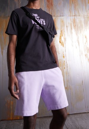 Mann trägt ein schwarzes Levi's T-Shirt, hellviolette Shorts und eine schwarze Umhängetasche vor einem rostigen Metallwand-Hintergrund.