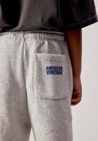 Personne portant un pantalon de survêtement gris clair avec une poche arrière étiquetée "AMERICAN VINTAGE" et un t-shirt noir ample.