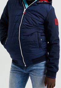 Námořnická pufovaná bunda vyrobená z prošívaného materiálu s kapucí, zipovým zapínáním a bočními kapsami. Obsahuje červený akcent na rukávu a žebrované manžety.