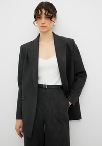 Blazer nero a righe sottili con vestibilità strutturata, abbinato a un top bianco e pantaloni a vita alta. Presenta una cintura nera e maniche extra-large.