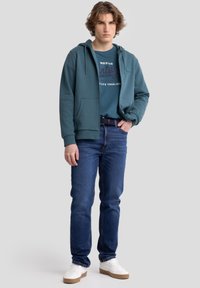 Heather krāsas kapuce ar priekšējām kabatām, pārklāta ar zilu grafisku T-kreklu, apvienota ar tumši ziliem džinsiem un baltiem sporta apaviem ar bežām zālēm.