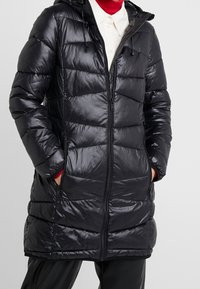 Vrouw in een lange zwarte gewatteerde pufferjas met een capuchon, rits aan de voorkant en handen in de zakken, over een witte shirt met kraag en een rode coltrui.