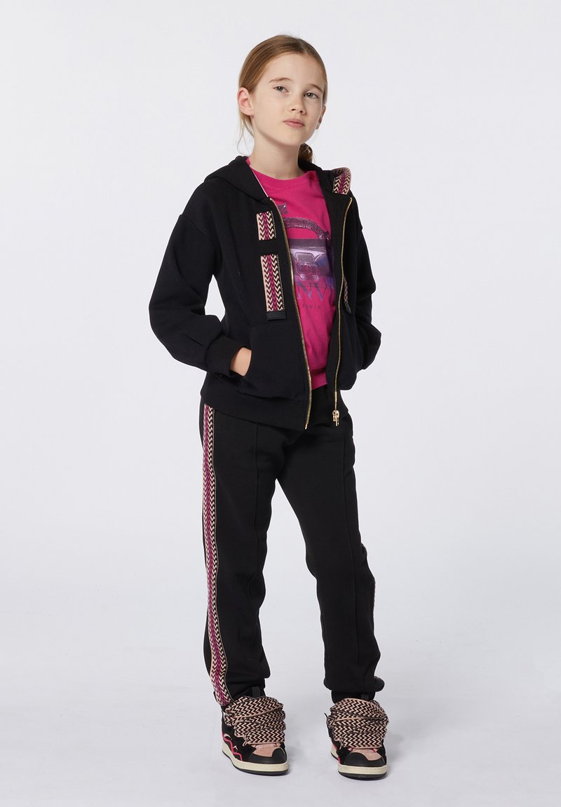 Sweat à capuche noir avec des accents à motifs, associé à un pantalon noir assorti avec des rayures latérales. T-shirt graphique rose en dessous. Baskets décontractées.