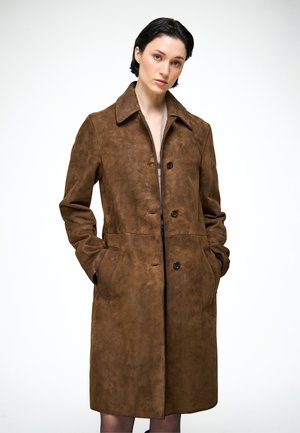 Femme aux cheveux courts et foncés portant un long manteau en suède marron avec des boutons, debout les mains dans les poches devant un fond blanc uni.