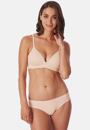 Femme aux cheveux brun mi-longs portant un soutien-gorge sans couture couleur chair et un sous-vêtement assorti, souriante et touchant ses cheveux.