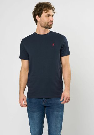 Donkerblauwe t-shirt met korte mouwen van katoen, voorzien van een klein rood geborduurd logo op de borst. Draagt met blauwe spijkerbroek.