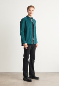 Camisa de botones en color verde azulado con cuello y tejido texturizado, utilizada sobre una camiseta negra, combinada con pantalones negros y zapatos negros. Acento de hebilla plateada.