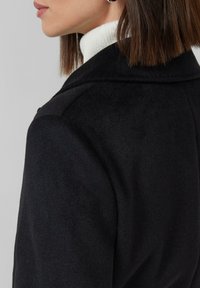 Cappotto in lana nera con colletto a punta, tessuto testurizzato e silhouette aderente, mostrato da dietro con un maglione a collo alto sotto.