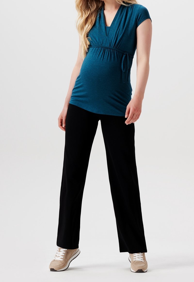 Sukienka ciążowa w kolorze teal z dekoltem w kształcie V, w talii empire i regulowanym zawiązaniu; w połączeniu z czarnymi spodniami o szerokich nogawkach i beżowymi sneakersami. Gładkie tekstury.