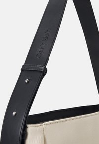 Anse de sac à main en cuir noir embossée "Calvin Klein" attachée au corps du sac en tissu beige.