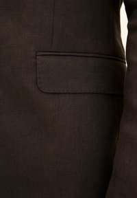 Un primo piano di un tessuto di blazer marrone scuro presenta una consistenza liscia, una tasca a toppa sul lato sinistro del petto e dettagli di cucitura sottili.