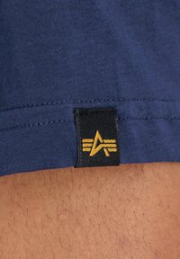 Marineblå bomullsskjorte med hem featuring et svart merke med en gul logo og tre horisontale linjer, synlige sømdetaljer.