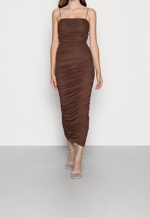 Kvinde iført en tætsiddende, rynket brun midi-kjole med tynde spaghetti-stropper og lyseblå, snørrede højhælede sandaler, stående på gråt gulv.