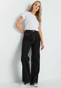 Jeans de pierna ancha negros con costuras blancas, cintura alta y cierre de botón, acompañados de una camiseta blanca sencilla y zapatillas negras.
