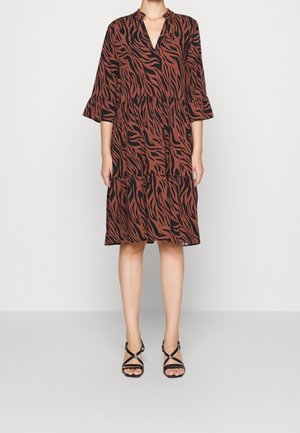 Kvinde iført en løs, knælængde brun kjole med sort zebraprint, trekvartærmer og sorte sandaler med remme mod en ensfarvet baggrund.