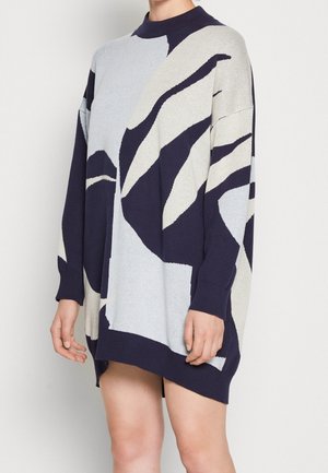 Kobieta w luźnej, długiej sukience-swetrze z abstrakcyjnymi czarno-biało-szarymi wzorami na tle jednolitego jasnego tła.