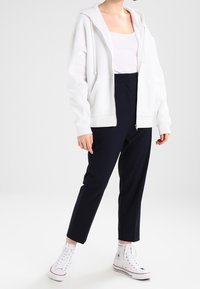 Haut à capuche zippé blanc avec capuche et cordons de serrage, porté par-dessus un débardeur léger, associé à un pantalon ajusté bleu marine et des baskets montantes blanches.