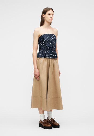 Γυναίκα με μπλε ναυτικό τοπ με πουά χωρίς τιράντες, μπεζ culottes με φαρδιά μπατζάκια, λευκές κάλτσες και καφέ πέδιλα με πλατφόρμα, στέκεται μπροστά σε απλό φόντο.