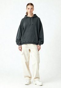 Felpa oversize grigia scura con tasca frontale e cordino, abbinata a pantaloni cargo color crema con tasche laterali e sneaker bianche.