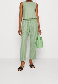 Tuta senza maniche verde con vita arricciata, abbinata a sandali neri con tacco. Una borsa verde coordinata completa il look.