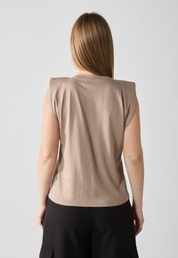 Top beige de manga corta con un material texturizado y un diseño estructurado en los hombros, combinado con pantalones negros, visto desde la parte de atrás.