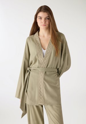 Giovane donna che indossa una giacca stile kimono larga verde oliva chiaro con cintura e pantaloni abbinati, in piedi davanti a uno sfondo chiaro e uniforme.