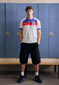 Maglia sportiva bianca con accenti rossi e blu, con l'emblema dell'Inghilterra, abbinata a pantaloni cargo neri e scarpe da ginnastica nere.