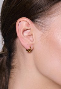 Złote kolczyki w kształcie obręczy, gładkie metalowe wykończenie, mały rozmiar, z wygiętym wzorem, który dodaje głębi i wymiaru, pokazane w zbliżeniu na uchu.