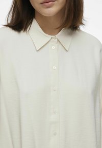 Camisa de botón de color crema claro con una textura suave, cuello estructurado y seis botones blancos en la parte delantera, con un corte relajado.