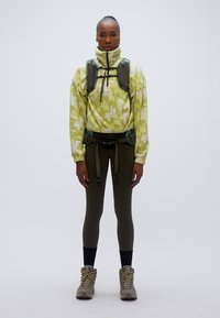 Φλις πουλόβερ σε ανοιχτό πράσινο καμουφλάζ, με μαύρο φερμουάρ, συνδυασμένο με ελιά κολάν και γκρι μπότες. Περιλαμβάνει μπροστινή ζώνη για προσάρτηση εξοπλισμού.