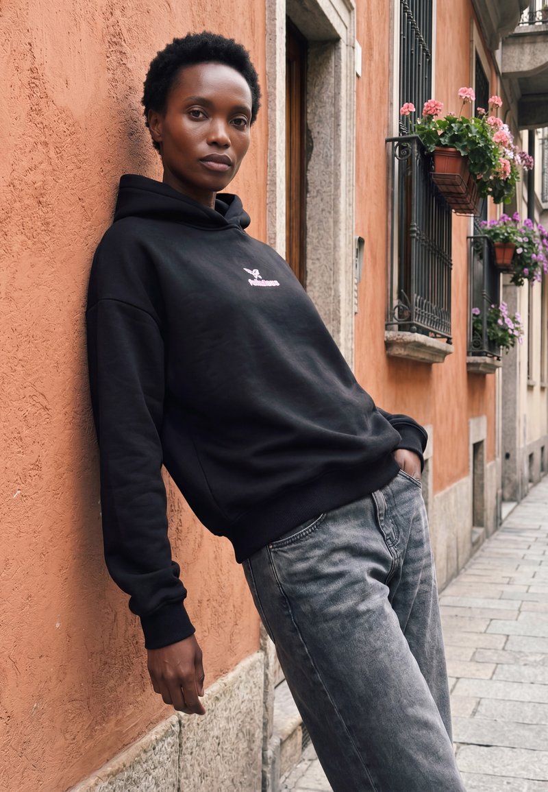 Frau mit kurzen lockigen Haaren lehnt an einer orangefarbenen Wand, trägt einen schwarzen Hoodie und graue Jeans in einer schmalen Straße mit Blumentöpfen an den Fenstern.