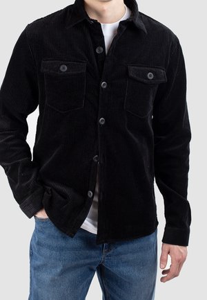 Mann trägt ein schwarzes Cordhemd über einem weißen T-Shirt, kombiniert mit blauen Jeans, die eine Hand in der Hosentasche hat.