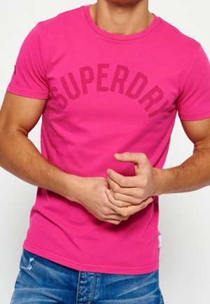 Mężczyzna w jaskraworóżowej koszulce Superdry i niebieskich dżinsach trzyma nadgarstek obiema rękami na jednolitym tle.