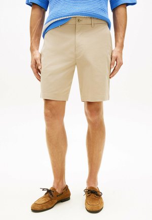 Homme portant un short beige mi-genou, un pull bleu texturé, et des mocassins en daim marron à lacets, debout devant un fond blanc.