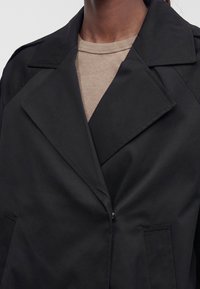 Gros plan d'une personne portant un trench noir sur un t-shirt beige côtelé à col rond, montrant le col et le haut du torse.