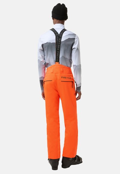 Jasnopomarańczowe spodnie śniegowe z regulowanymi czarnymi szelkami, kieszeniami na zamek i gładką fakturą, noszone na wzorzystej koszuli z długim rękawem.