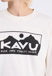 Weißes Langarmshirt mit einem großen schwarzen Grafikdesign, das Berge, eine Sonne und den Text "KAVU KLEAR ABOVE VISIBILITY UNLIMITED" zeigt.