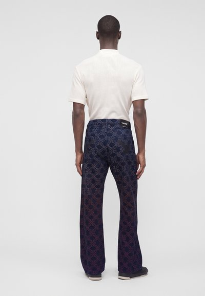 Homme debout, de dos, portant une chemise blanche texturée à manches courtes et un pantalon large à motifs sombres avec des chaussures noires.
