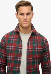 Rood en groen geblokte flanellen shirt met een knoopsluiting aan de voorkant en een borstzak, met een logo patch en een zachte textuur. Grijze basislaag eronder.