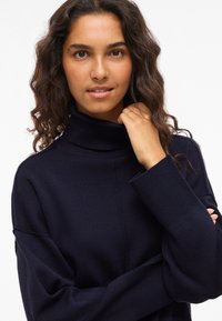 Marineblauer Strick-Turtleneck-Pullover mit weiten Ärmeln und gerippten Bündchen, der eine glatte Textur und eine lockere Passform aufweist und lässig getragen wird.